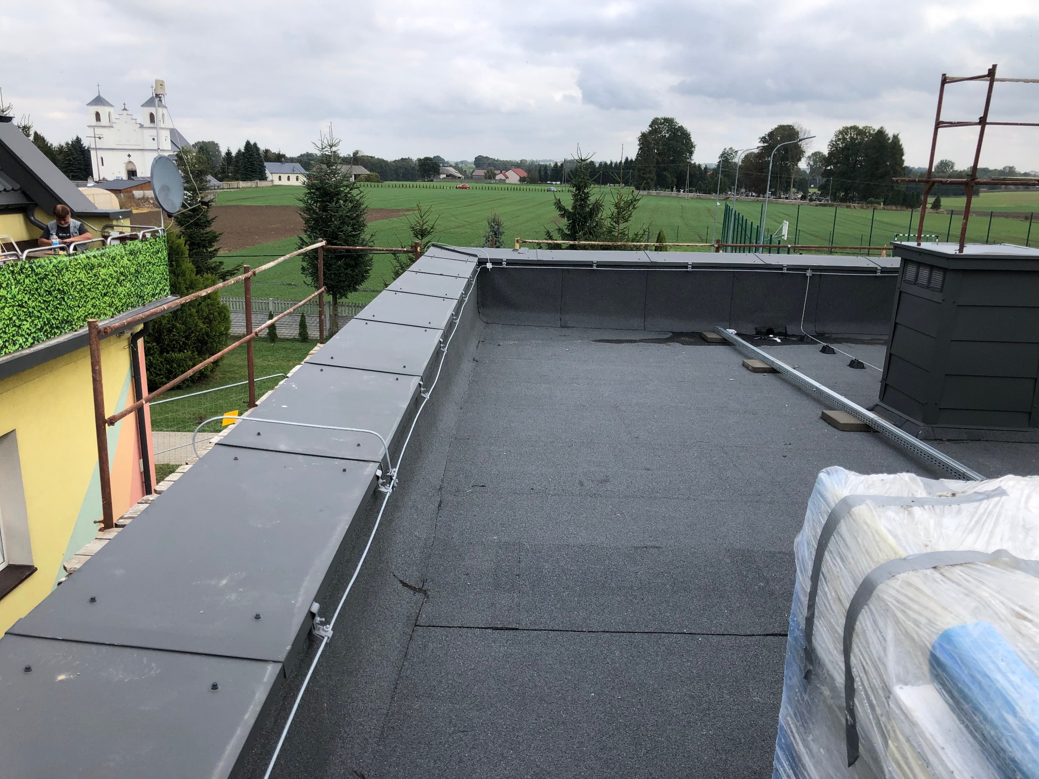 Szary dach płaski z zamontowanymi elementami instalacji odgromowej, widok na okoliczne pola i budynki w pochmurny dzień.