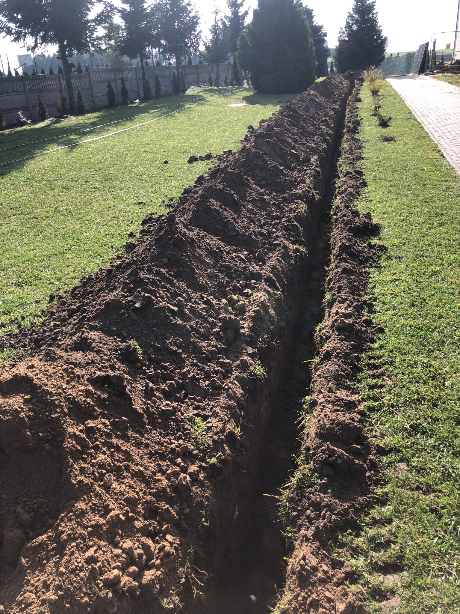 Wykopany głęboki rów w trawniku, prawdopodobnie pod kable lub rury, z pryzmami ziemi po obu stronach, widok z góry w słoneczny dzień.