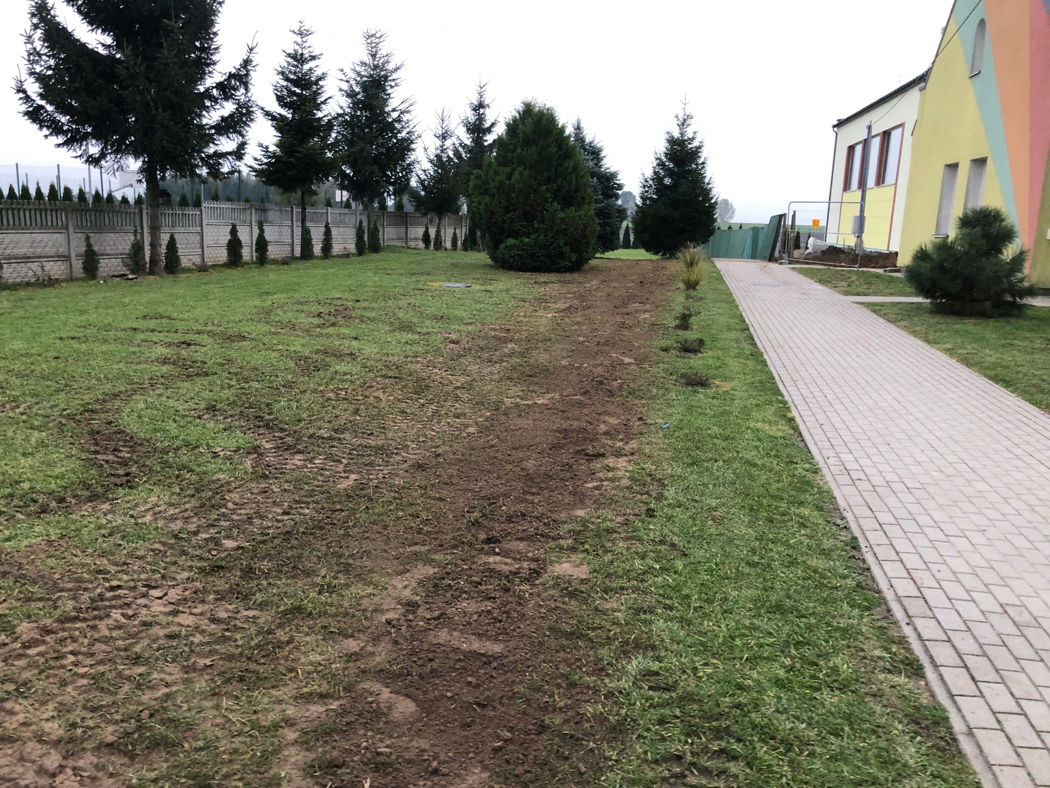 Teren zielony z widocznymi śladami po pracach ziemnych, prawdopodobnie przygotowany pod instalację odgromową, w tle ogrodzenie i budynek z kolorową elewacją.