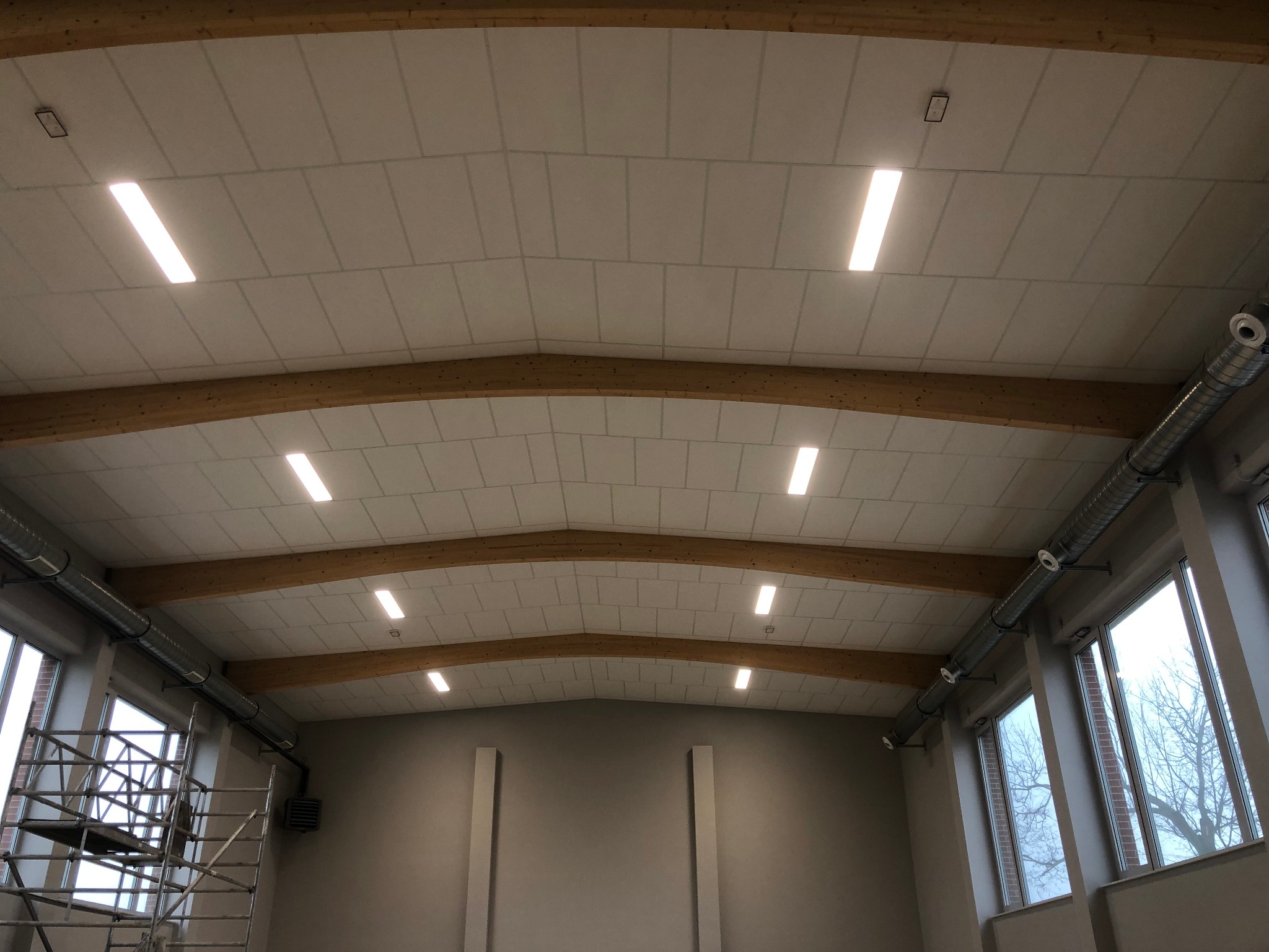 Wnętrze hali sportowej z widocznym sufitem podwieszanym z oświetleniem ledowym, drewnianymi belkami stropowymi, systemem wentylacji i rusztowaniem przy oknie.