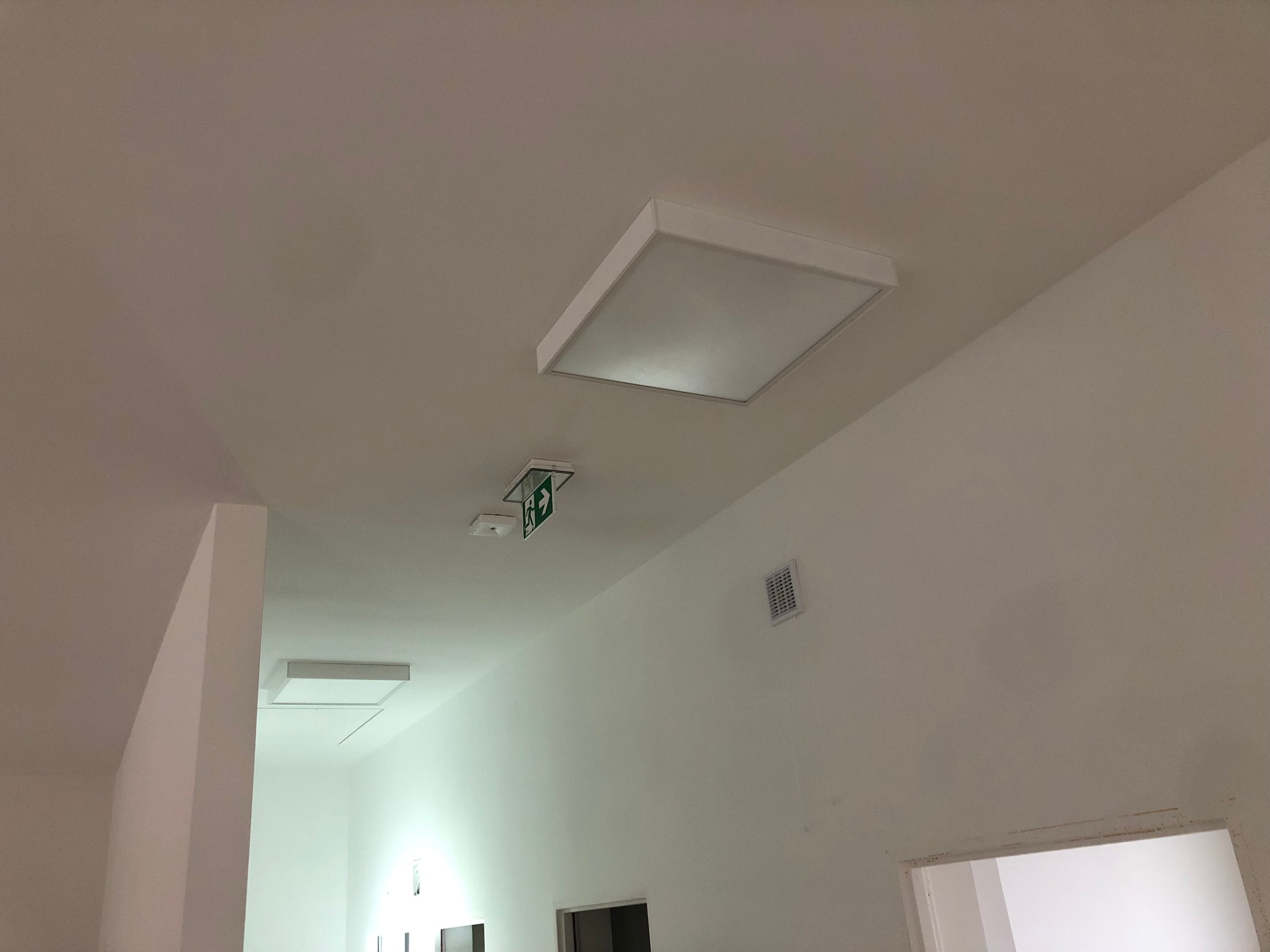 Białe, kwadratowe oprawy oświetleniowe LED zamontowane na suficie w korytarzu biurowym, widoczny znak ewakuacyjny i kratka wentylacyjna na ścianie.