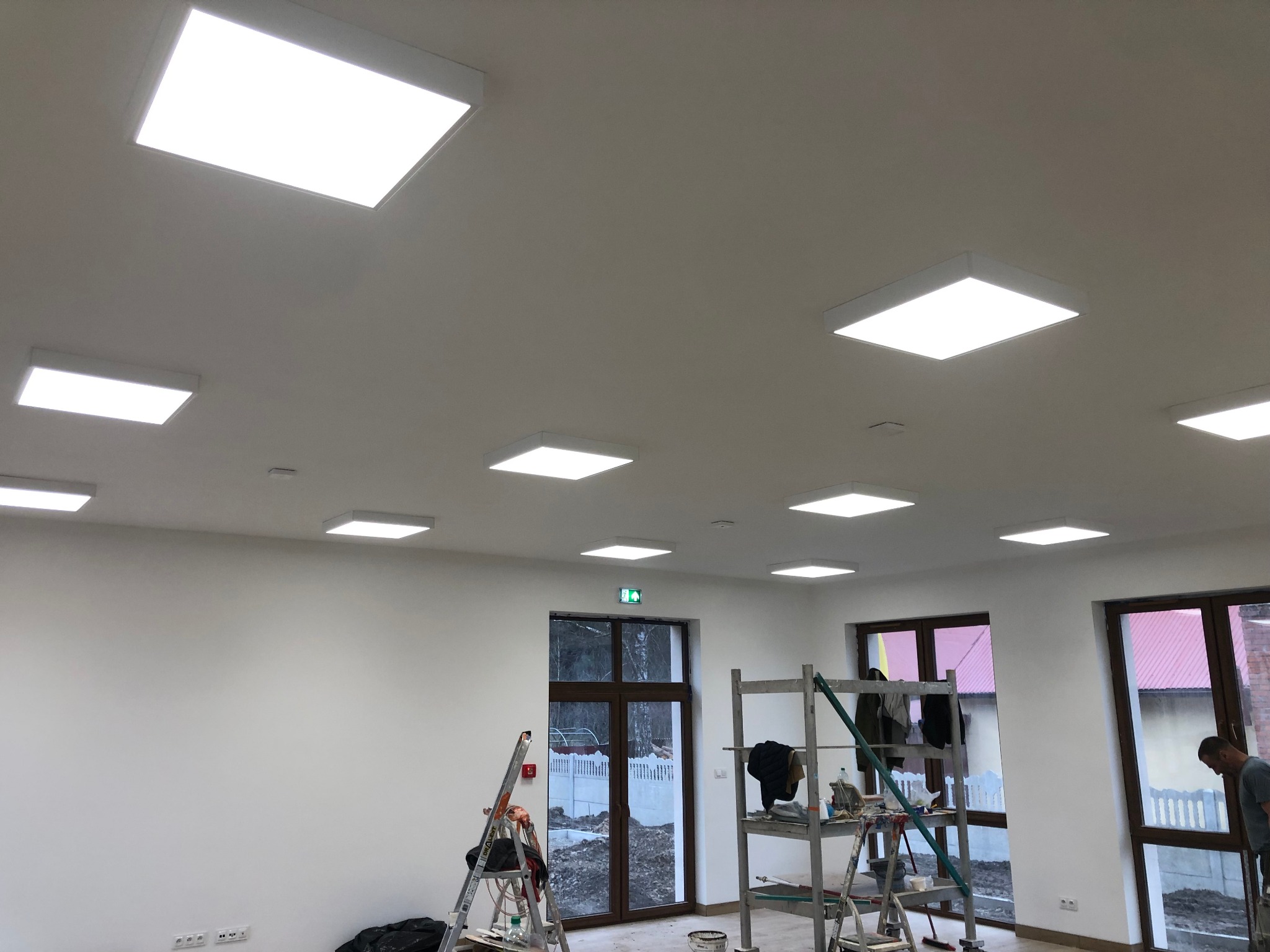 Montaż paneli LED w jasnym pomieszczeniu z białymi ścianami, widoczne drabiny i rusztowania, okna z brązowymi ramami, częściowo widoczna osoba w prawym dolnym rogu.