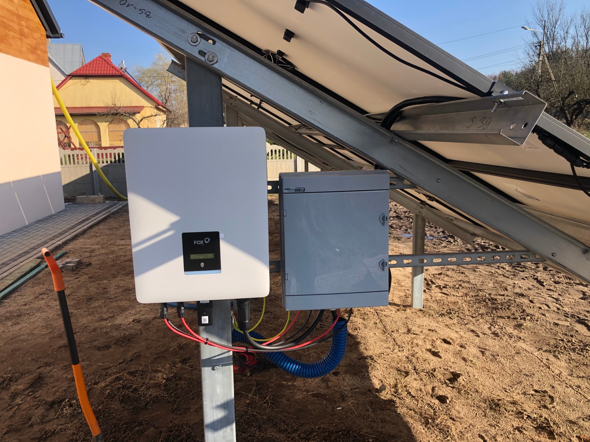 Instalacja paneli fotowoltaicznych: widok na falownik Fox ESS, skrzynkę przyłączeniową, kable w niebieskiej karbowanej osłonie, oraz fragment paneli na metalowej konstrukcji, z widoczną łopatą w tle.