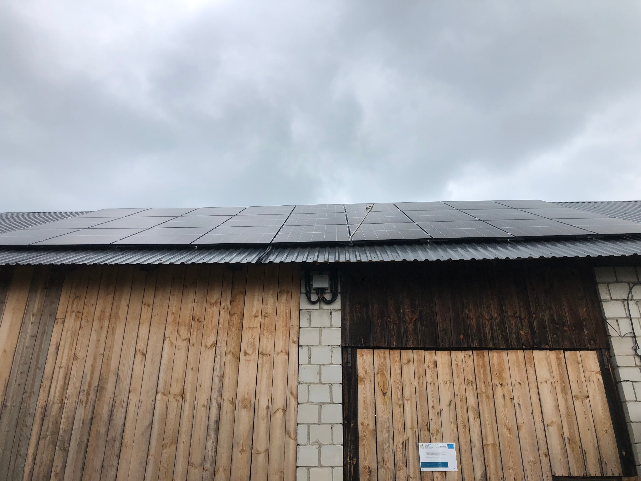Panele fotowoltaiczne na dachu budynku gospodarczego z drewnianymi wrotami, widoczna skrzynka elektryczna i dokumentacja techniczna na ścianie.