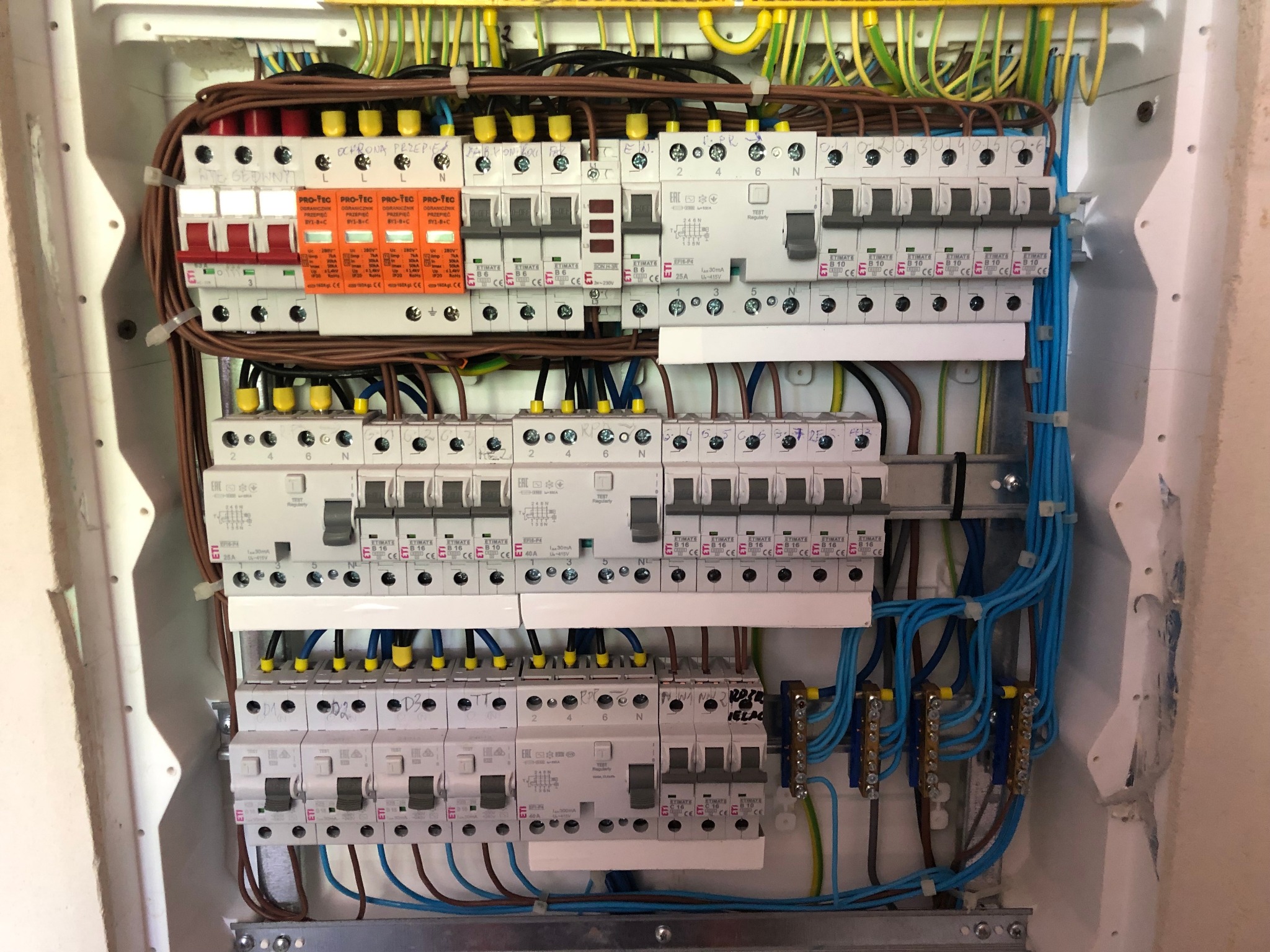 Rozdzielnica elektryczna z zabezpieczeniami nadprądowymi, ogranicznikami przepięć i przewodami w różnych kolorach, zamontowana wewnątrz obudowy z widocznymi śladami napraw na ścianie.
