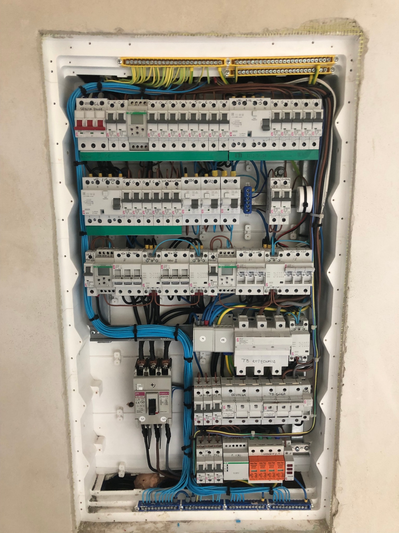 Rozdzielnia elektryczna z zainstalowanymi bezpiecznikami, przewodami i listwami zaciskowymi w białej obudowie, widoczne oznaczenia na elementach i przewody w kolorach niebieskim, żółto-zielonym...
