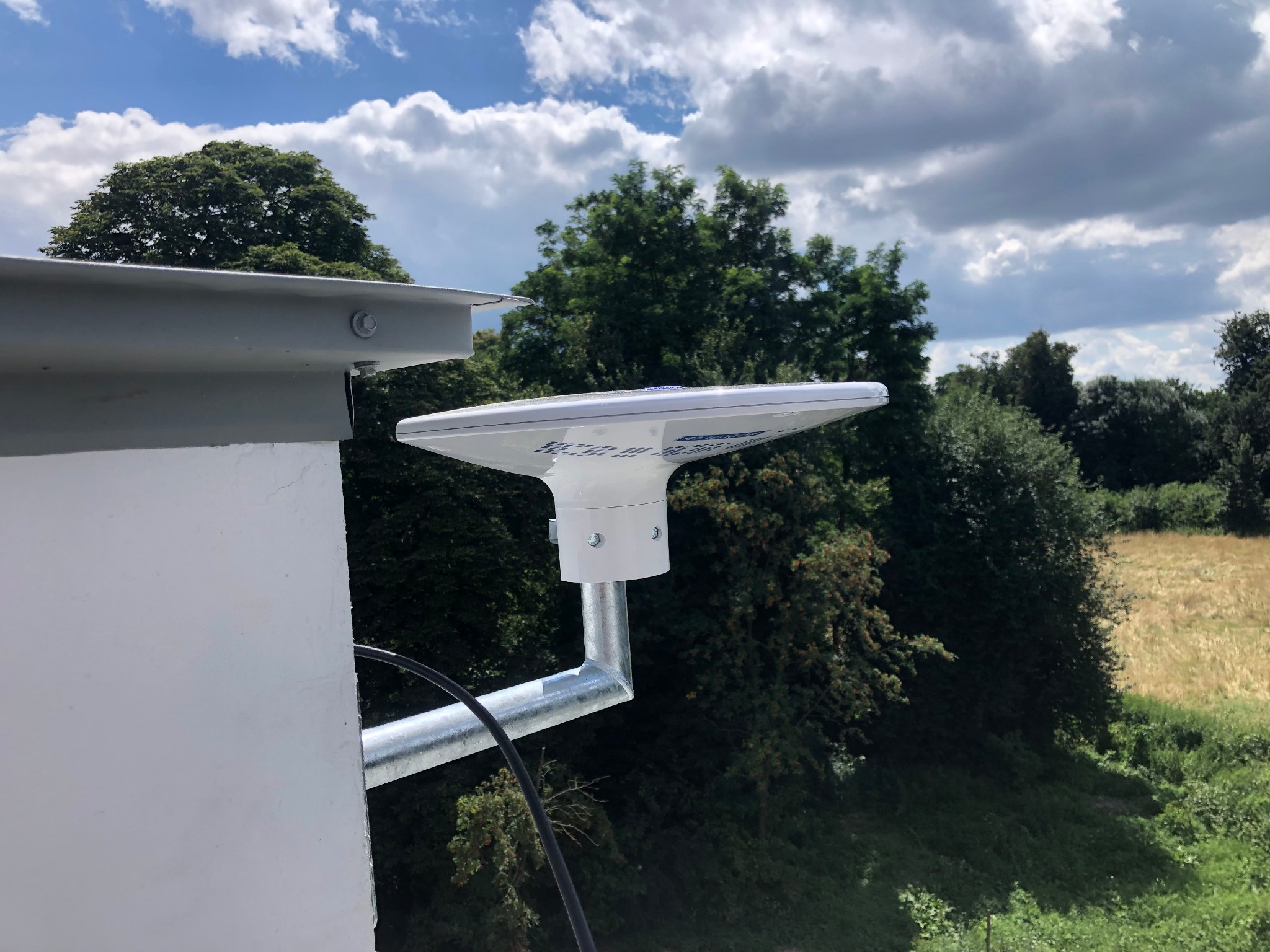 Zewnętrzna antena zamontowana na budynku, widok z dołu na tle drzew i częściowo zachmurzonego nieba.