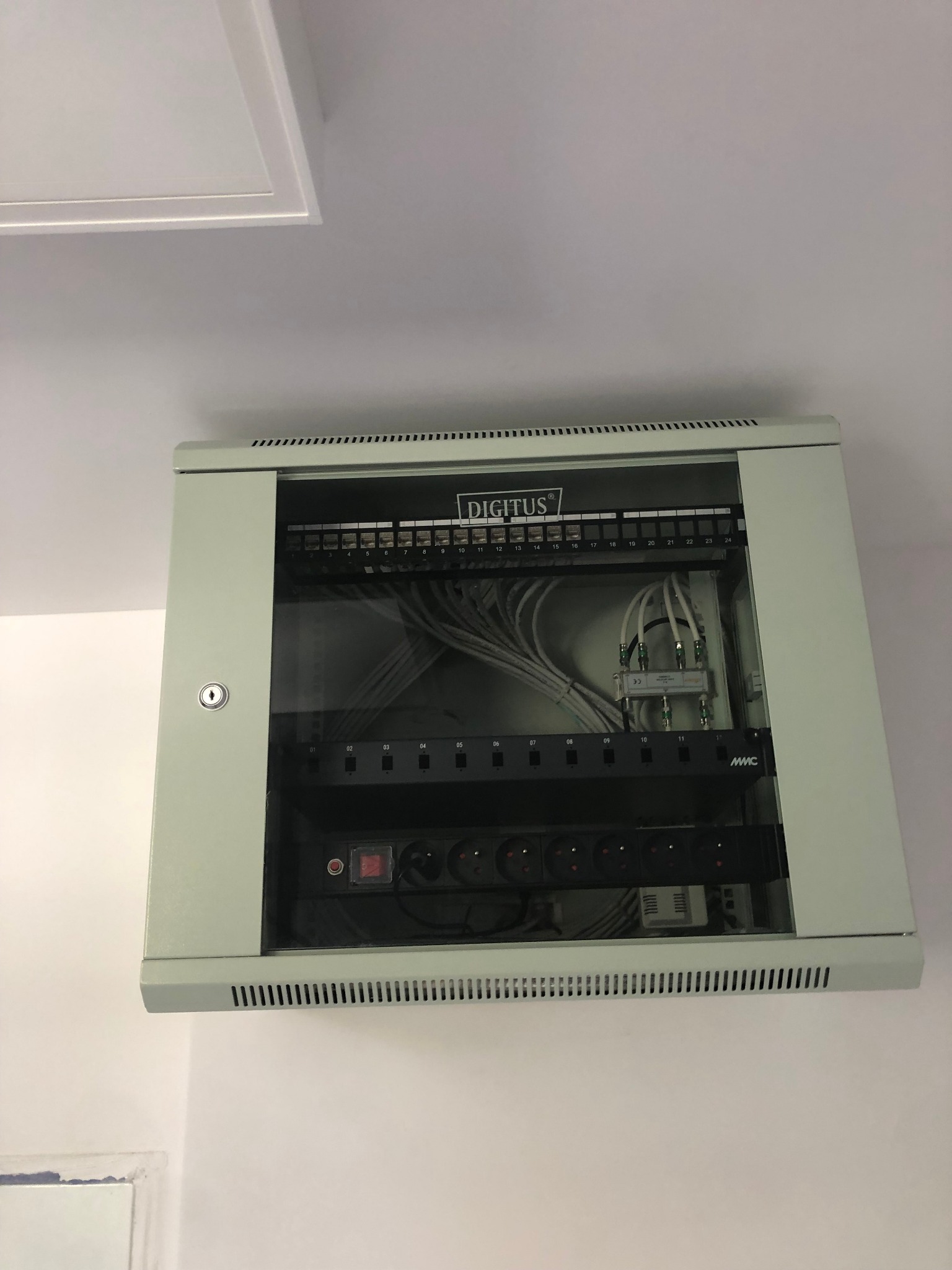 Zamknięta szafa rack DIGITUS z widocznymi kablami, rozdzielaczami i listwą zasilającą, zamontowana na białej ścianie pod sufitem.