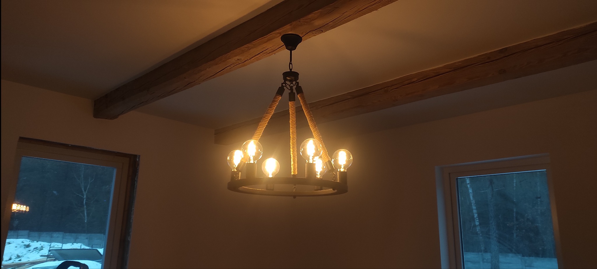Rustykalny żyrandol z żarówkami Edisona zawieszony na sznurach, widoczne drewniane belki stropowe i okno z widokiem na zimowy krajobraz.