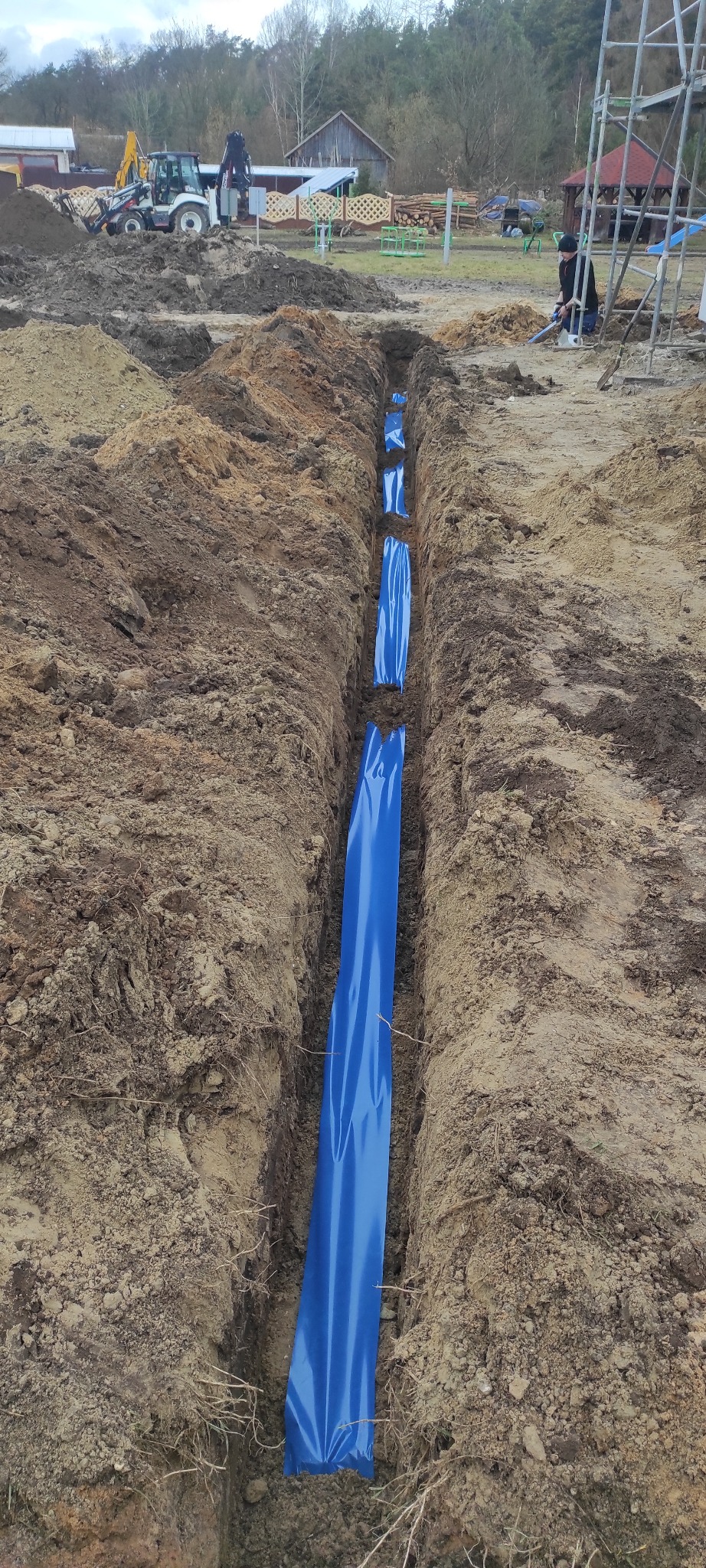 Wykop w ziemi, wyłożony niebieską folią ochronną, przygotowany pod instalację elektryczną lub rury, w tle koparka i elementy konstrukcji.