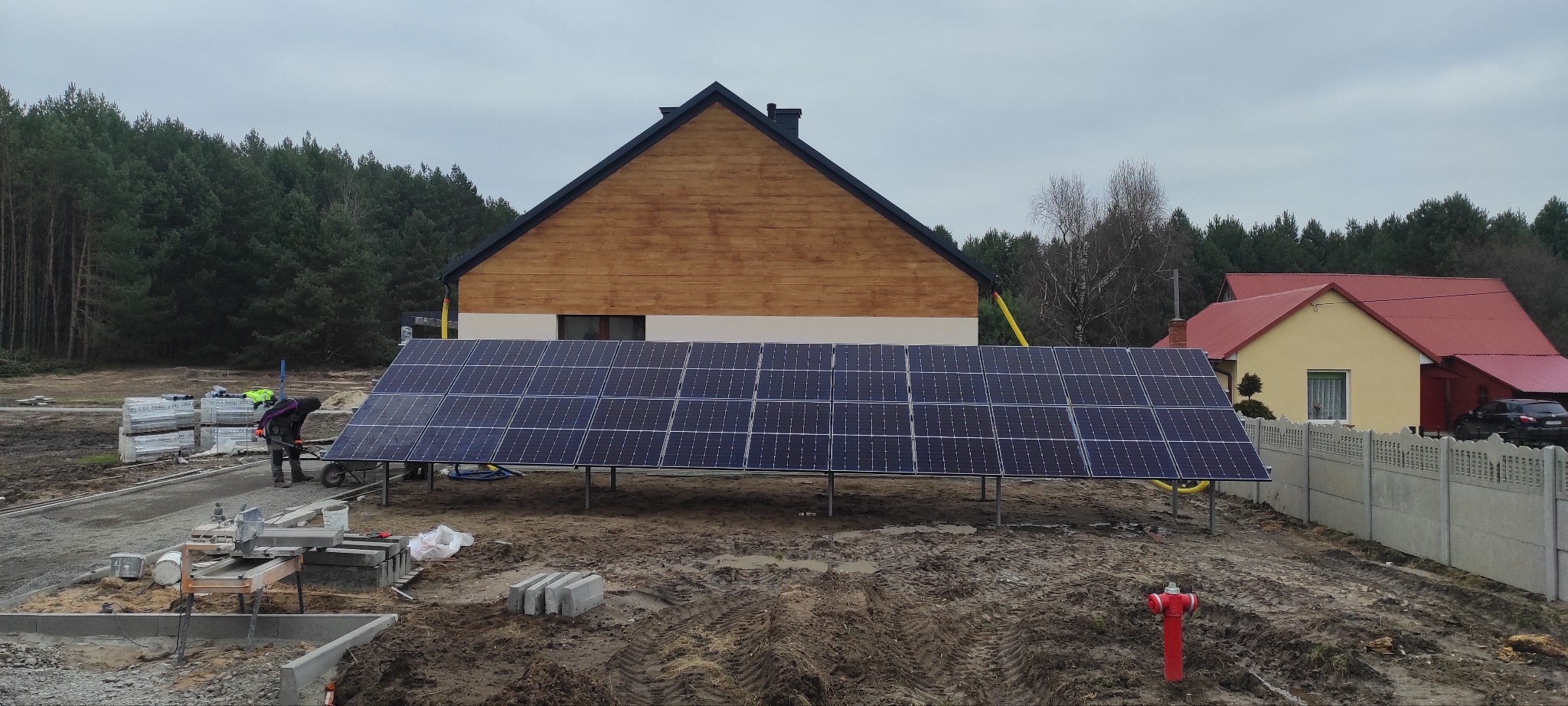 Montaż paneli fotowoltaicznych na gruncie przed domem jednorodzinnym, widoczny pracownik w odzieży roboczej i narzędzia budowlane, w tle las i budynki.