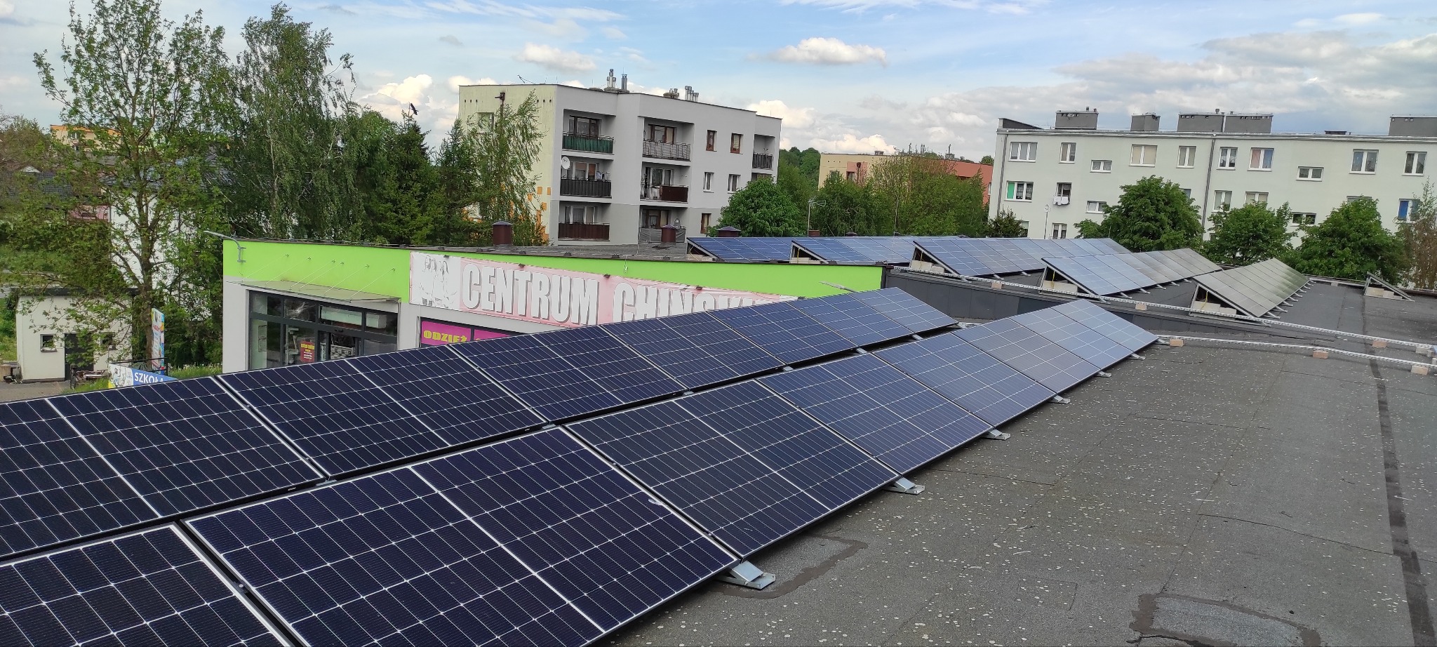 Rząd paneli fotowoltaicznych na dachu budynku handlowego z zielonym dachem, widok z góry na tle zabudowy miejskiej i błękitnego nieba z chmurami.