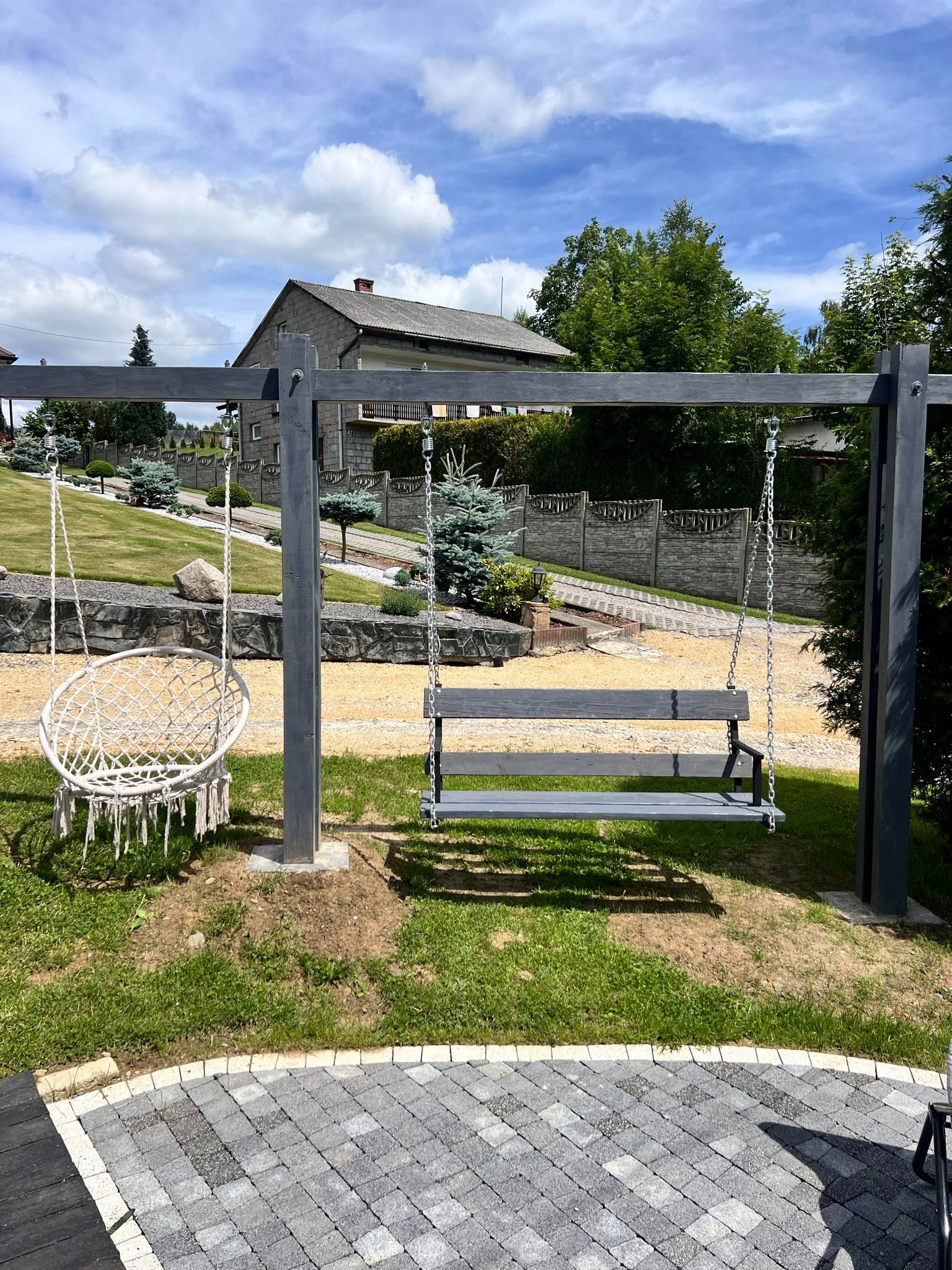Nowoczesny plac zabaw z huśtawkami na metalowym stelażu, wykończony kostką brukową w odcieniach szarości, na tle zadbanego ogrodu z domem.