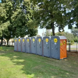 Szereg szarych przenośnych toalet WC SKIP ustawionych na trawniku pod drzewami w słoneczny dzień. Jedna toaleta ma pomarańczowe elementy.