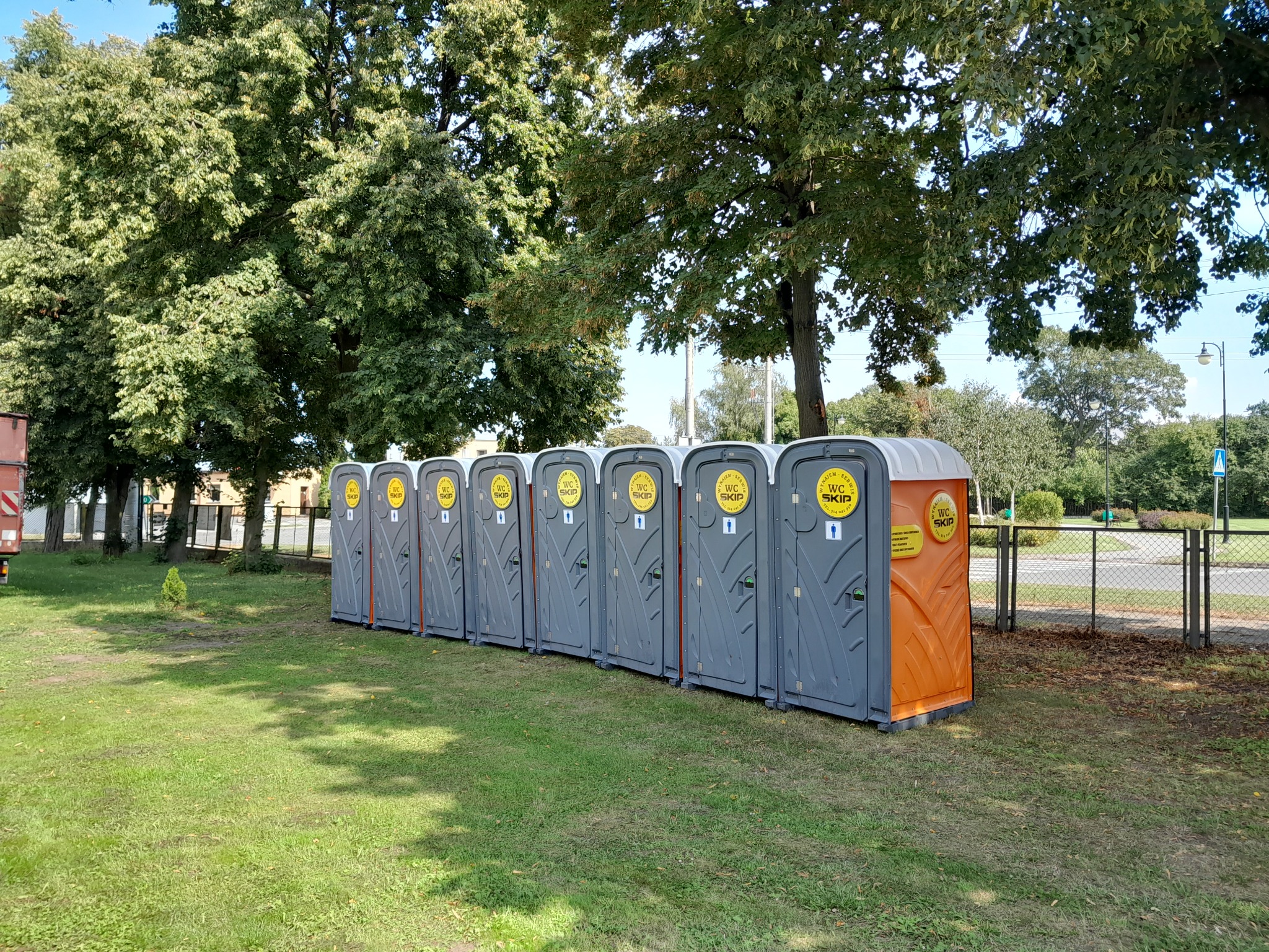 Szereg szarych przenośnych toalet WC SKIP ustawionych na trawniku pod drzewami w słoneczny dzień. Jedna toaleta ma pomarańczowe elementy.
