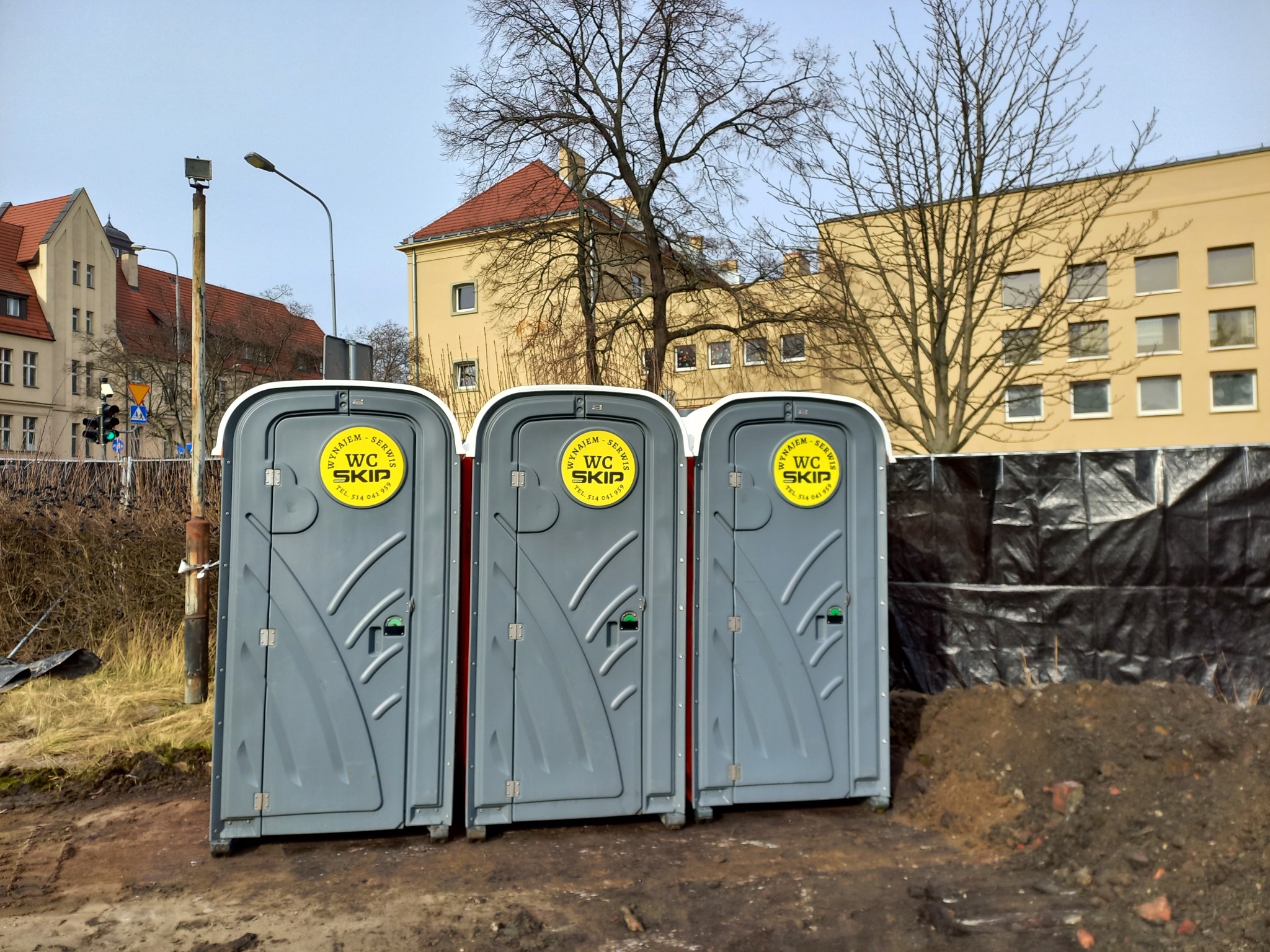 Trzy przenośne toalety WC SKIP ustawione obok siebie na placu budowy, widoczne budynki w tle, ponura pogoda.