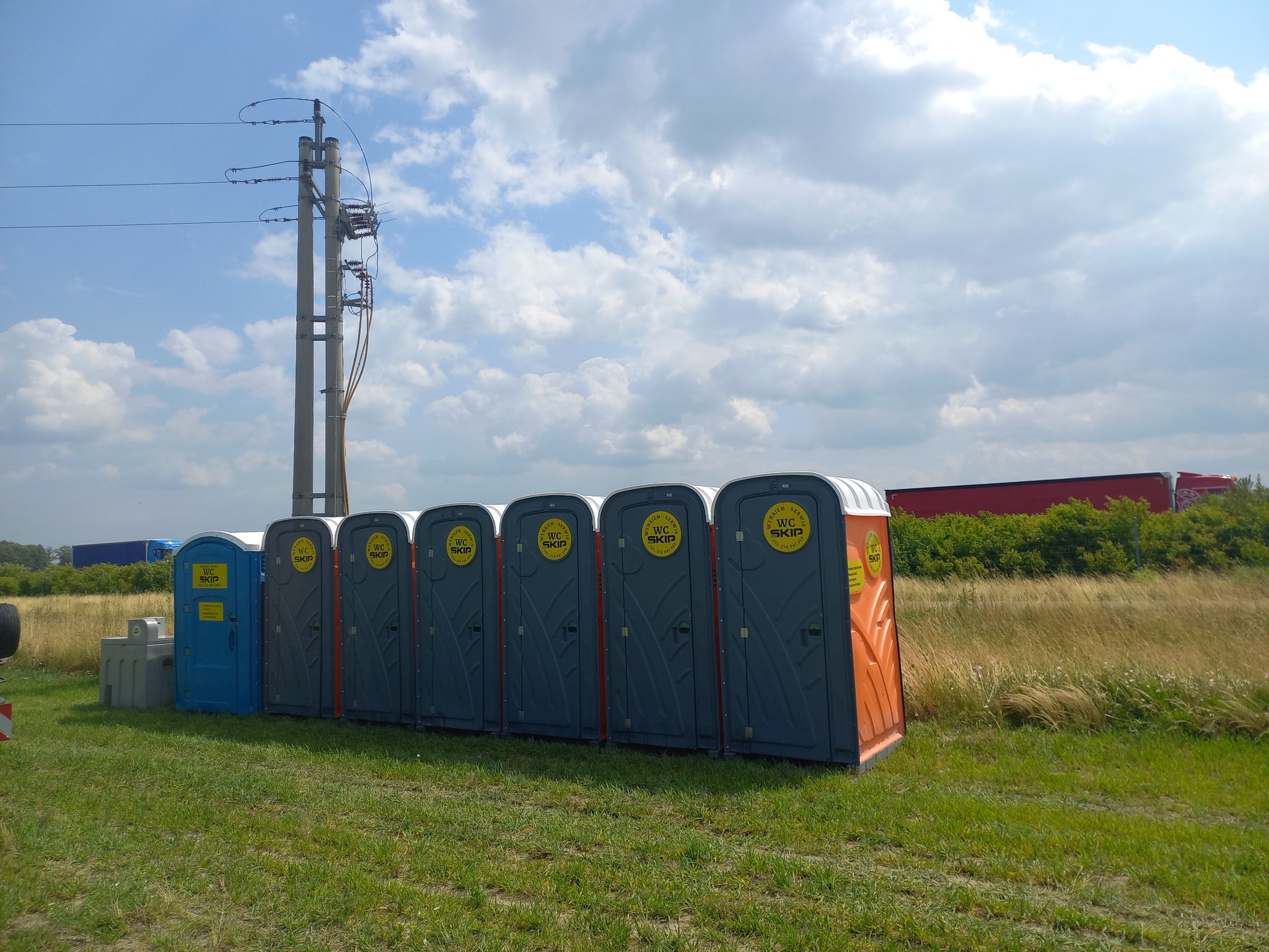 Rząd przenośnych toalet WC SKIP w różnych kolorach (niebieski, szary, pomarańczowy) ustawionych na trawiastym terenie, z widoczną linią wysokiego napięcia i przejeżdżającymi pojazdami w tle.