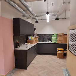 Usługi Remontowo-Wykończeniowe Dawid Zając - Wnętrze kuchni w Szczecinie w trakcie wykańczania, widoczne ciemne szafki, biały blat, ułożona podłoga w jodełkę, kartony i butelka wina na blacie, oraz rośliny doniczkowe.