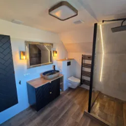 Nowoczesna łazienka na poddaszu z drewnopodobnymi płytkami, czarną armaturą, umywalką nablatową w odcieniach granatu i złota oraz kabiną prysznicową typu walk-in z podświetleniem LED.