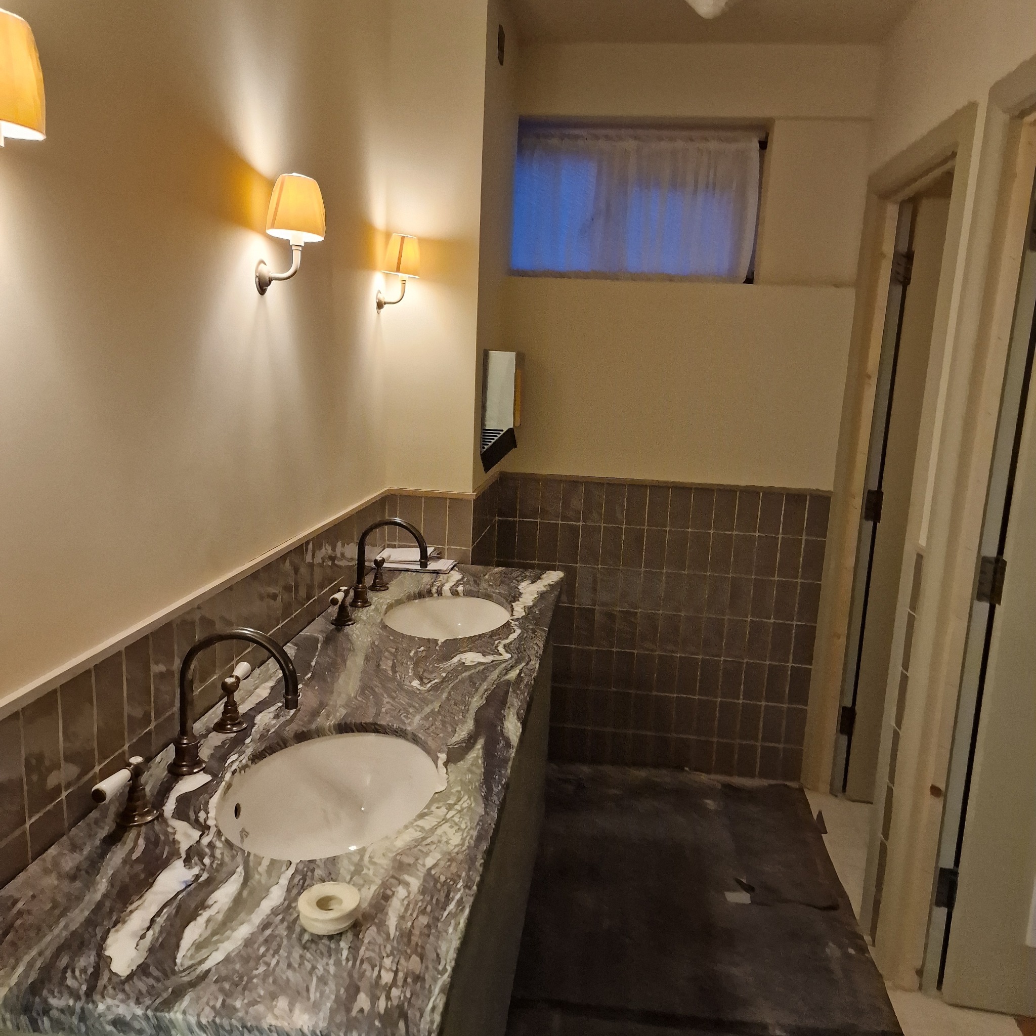 Łazienka w trakcie wykończenia: marmurowy blat z dwoma umywalkami i bateriami w stylu retro, oświetlona trzema kinkietami z abażurami, widoczne niedokończone podłogi i ościeżnice drzwi.