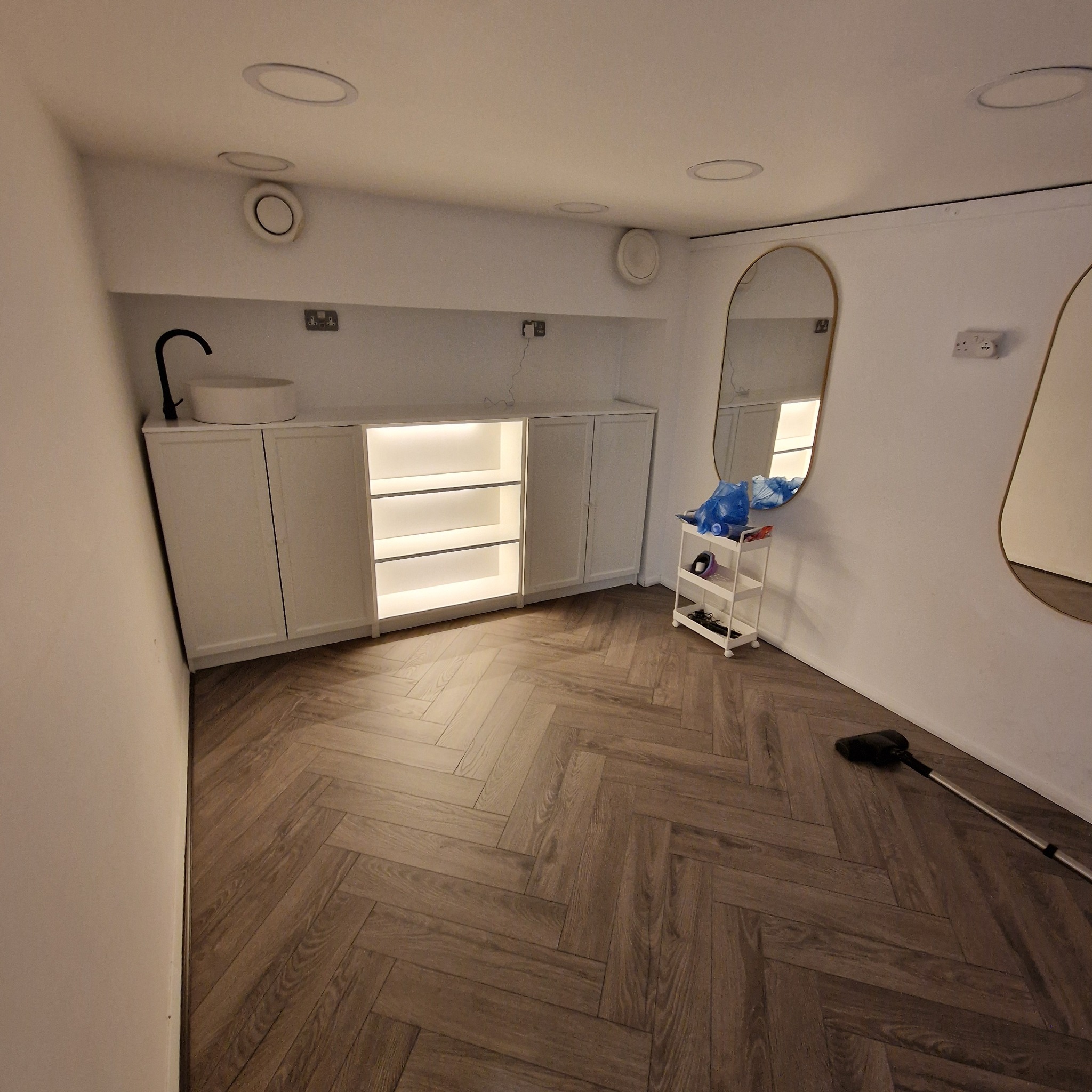 Nowoczesna łazienka z szafkami pod umywalkę z podświetlanymi półkami, dwoma owalnymi lustrami z cienką złotą ramą, podłogą ułożoną w jodełkę, plastikowym wózkiem z przyborami i odkurzaczem w rogu.