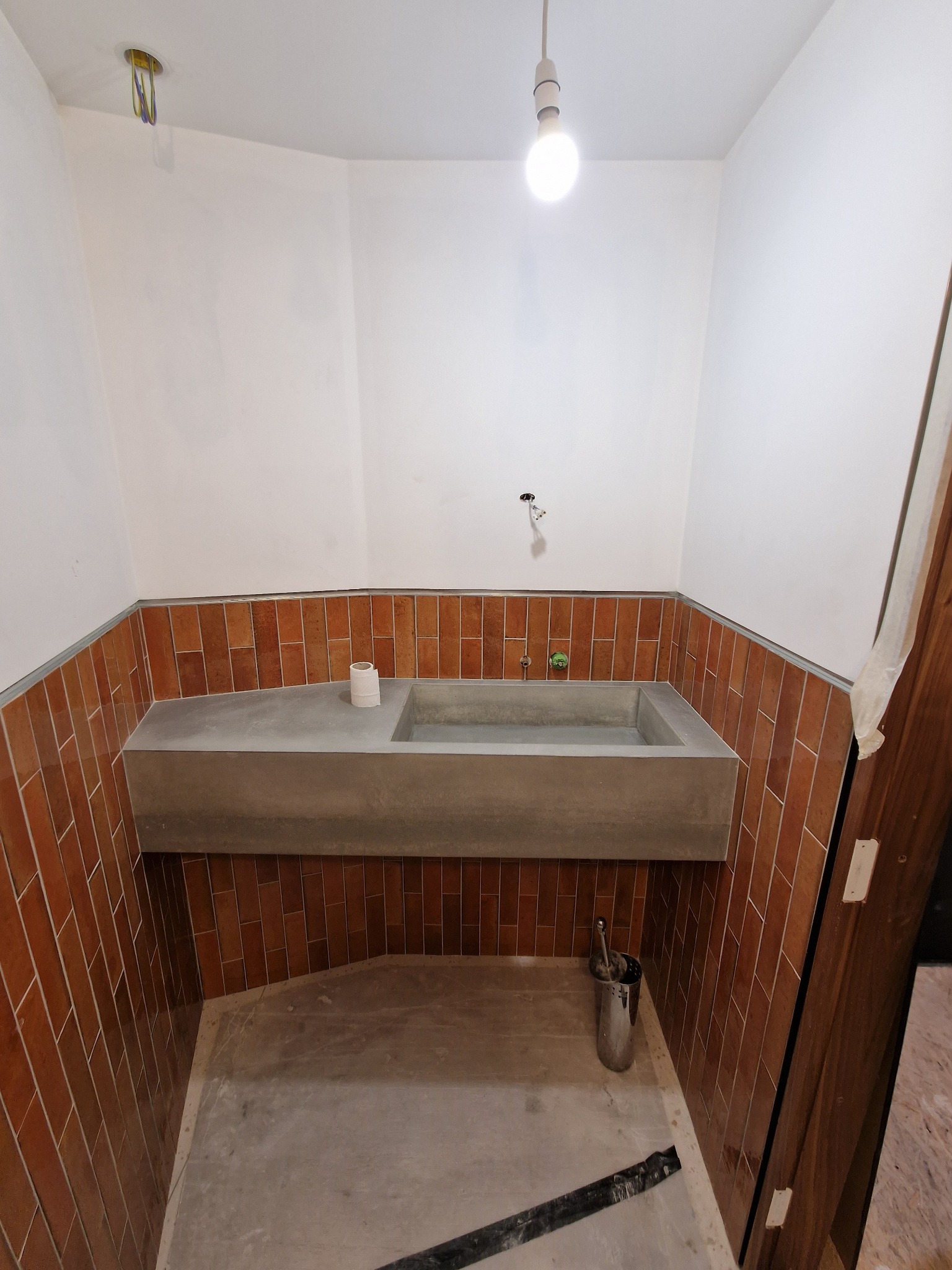 Łazienka w trakcie wykańczania: surowy betonowy blat z umywalką, brązowe płytki na ścianach, widoczne niedociągnięcia i narzędzia, surowy stan podłogi, żarówka na kablu.