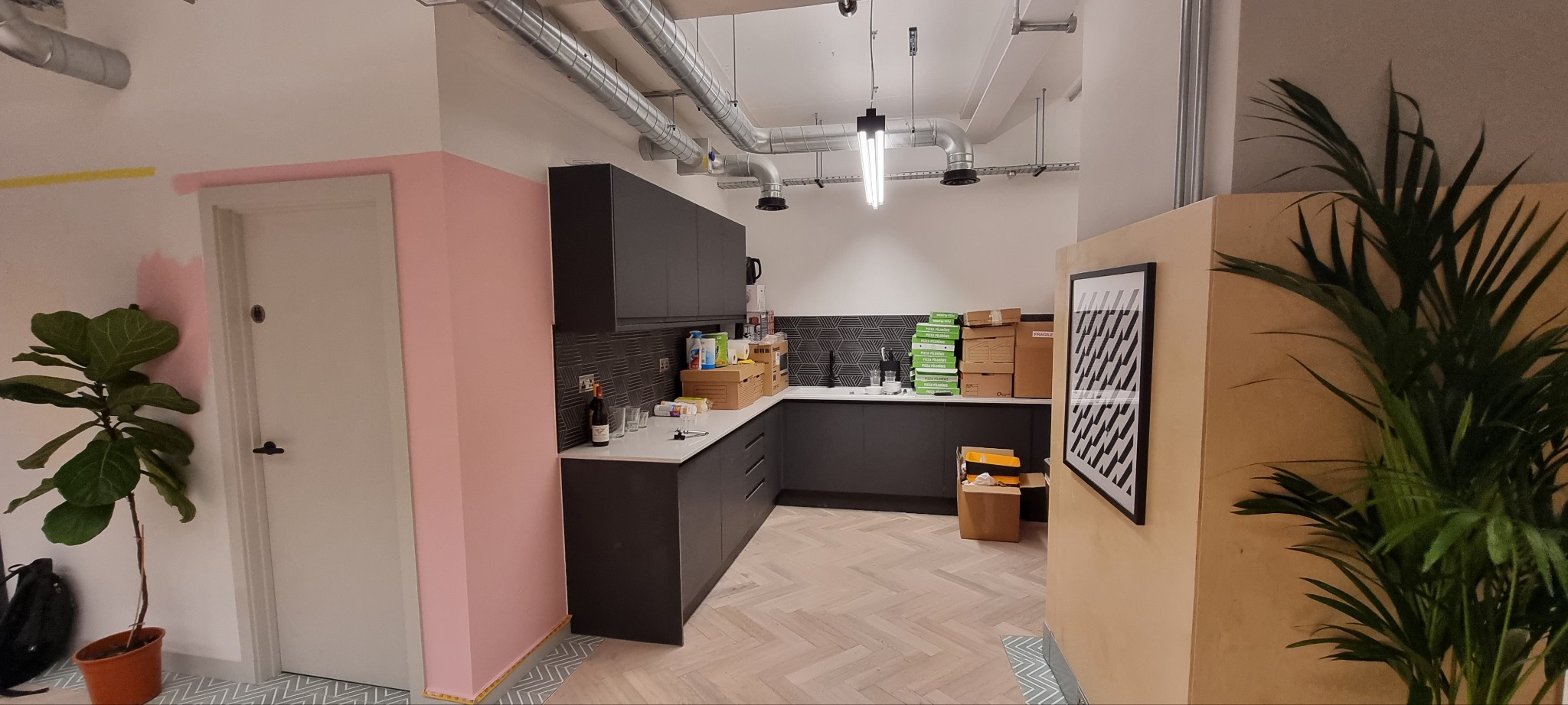 Wnętrze kuchni w Szczecinie w trakcie wykańczania, widoczne ciemne szafki, biały blat, ułożona podłoga w jodełkę, kartony i butelka wina na blacie, oraz rośliny doniczkowe.