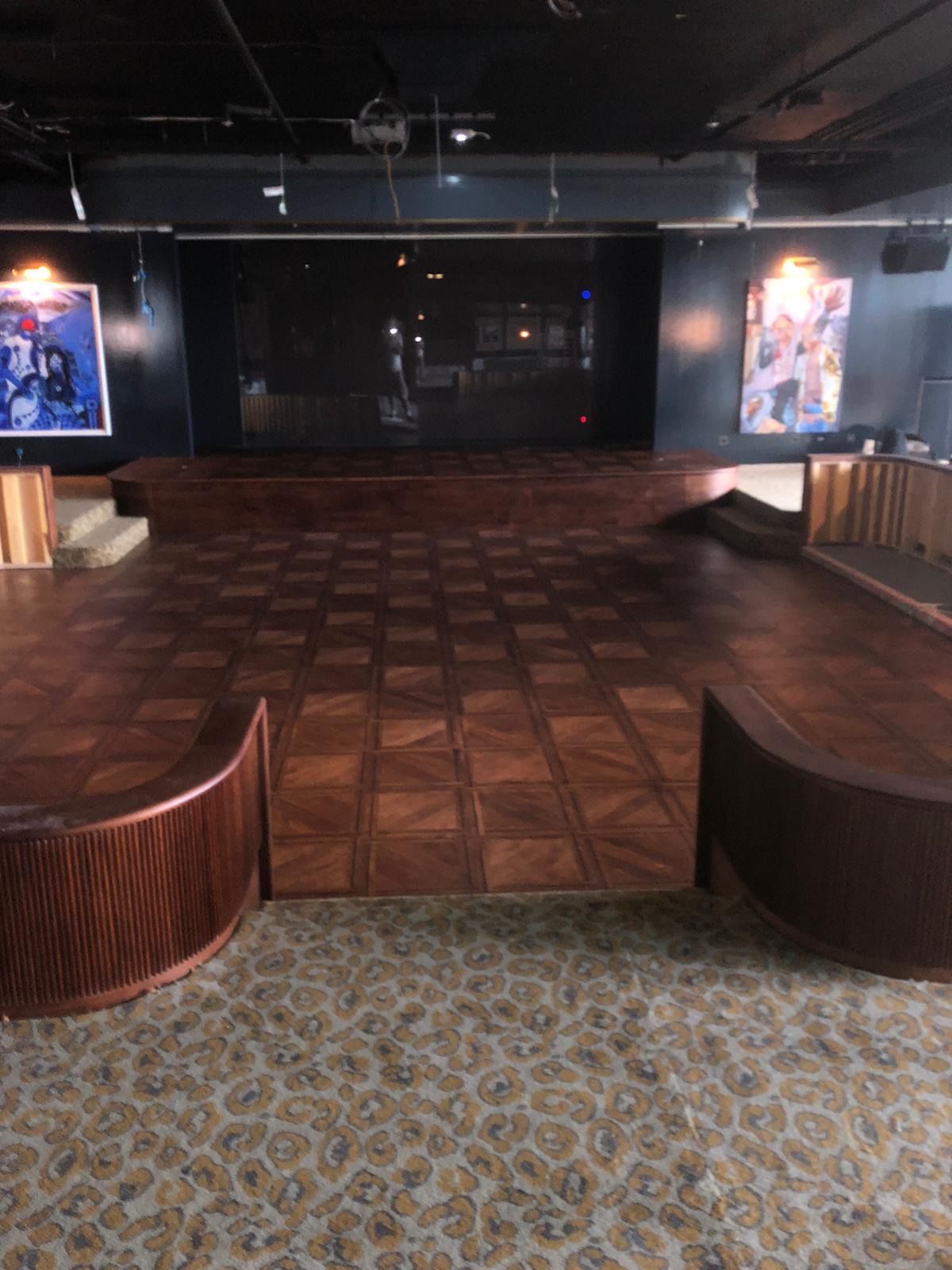 Sala z parkietem ułożonym w kwadraty, podwyższona scena w tle i dywan w panterkę na pierwszym planie.