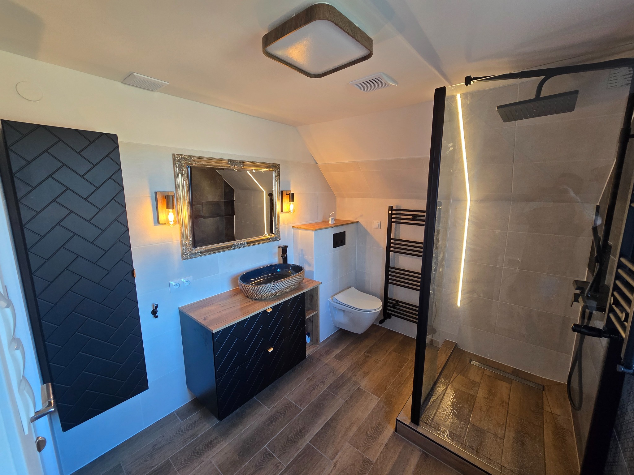 Nowoczesna łazienka na poddaszu z drewnopodobnymi płytkami, czarną armaturą, umywalką nablatową w odcieniach granatu i złota oraz kabiną prysznicową typu walk-in z podświetleniem LED.