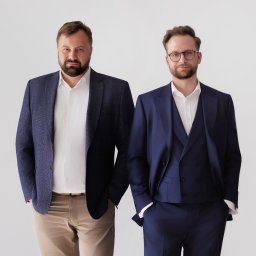 5E Spółka Doradztwa Podatkowego - Dwóch eleganckich mężczyzn w biznesowych strojach: jeden w granatowej marynarce i beżowych spodniach, drugi w trzyczęściowym garniturze i okularach, stoi na jasnym tle, patrząc wprost w obiektyw.