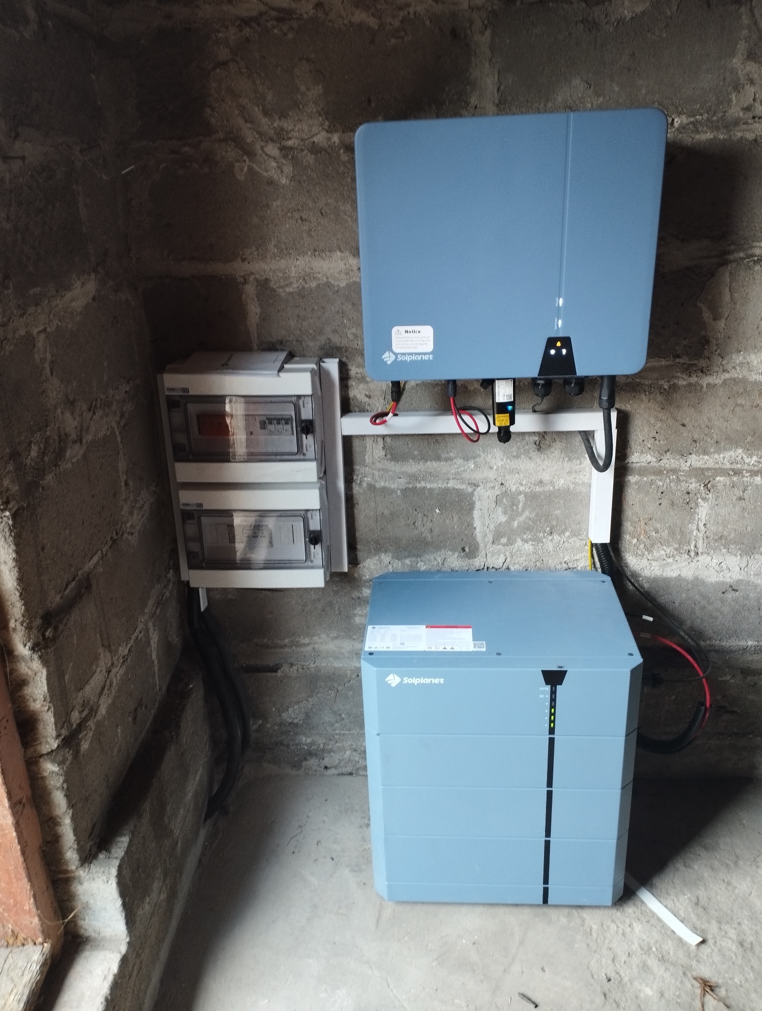 Zainstalowany magazyn energii Solplanet wraz z inwerterem i rozdzielnicą elektryczną na tle surowej ściany z cegły, wewnątrz budynku. Widoczne okablowanie.