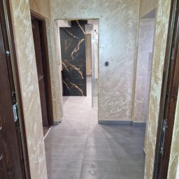 Dzik-Rem - Korytarz po remoncie z szarymi płytkami podłogowymi, ścianami wykończonymi dekoracyjną techniką malarską w odcieniach beżu i złotymi akcentami, widok na łazienkę z czarnymi płytkami imitującymi...