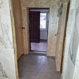 Dzik-Rem - Korytarz po remoncie: szare płytki podłogowe, ściany z efektem dekoracyjnym w odcieniach beżu i brązu, marmurowe wykończenie ścian po lewej stronie, widoczne niedociągnięcia w wykończeniu framugi...