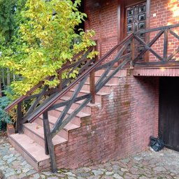Dzik-Rem - Zewnętrzne schody wejściowe z brązowych płytek i drewnianą balustradą przy domu z czerwonej cegły, widoczne ślady zabrudzeń na cegłach, obok rośnie drzewo z żółtymi liśćmi, na dole zwinięty kabel.