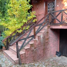 Zewnętrzne schody wejściowe z brązowych płytek i drewnianą balustradą przy domu z czerwonej cegły, widoczne ślady zabrudzeń na cegłach, obok rośnie drzewo z żółtymi liśćmi, na dole zwinięty kabel.