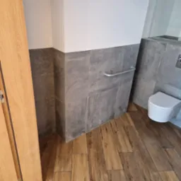 Wykończona łazienka z drewnopodobną podłogą, szarymi płytkami do połowy ściany i białą miską WC. Widoczny fragment drzwi. Nowoczesny design.