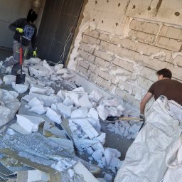 Dzik-Rem - Demontaż ocieplenia: Dwóch robotników usuwa styropian ze ściany, przygotowując podłoże. Fragmenty izolacji i narzędzia na podłodze.