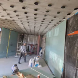 Montaż płyt karton-gips na ścianie i suficie z widocznymi elementami mocującymi. Pracownik na drabinie, narzędzia i materiały budowlane w pomieszczeniu w trakcie prac.