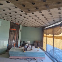 Dzik-Rem - Montaż płyt karton-gips na suficie. Widoczne płyty, narzędzia pracy, drabina, okno. Sufit z elementami mocującymi. Prace wykończeniowe w jasnym pomieszczeniu.