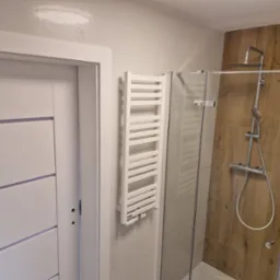 Nowoczesna łazienka po remoncie: białe drzwi z poziomymi frezami, grzejnik drabinkowy i kabina prysznicowa z drewnopodobnymi płytkami. Minimalistyczny design.
