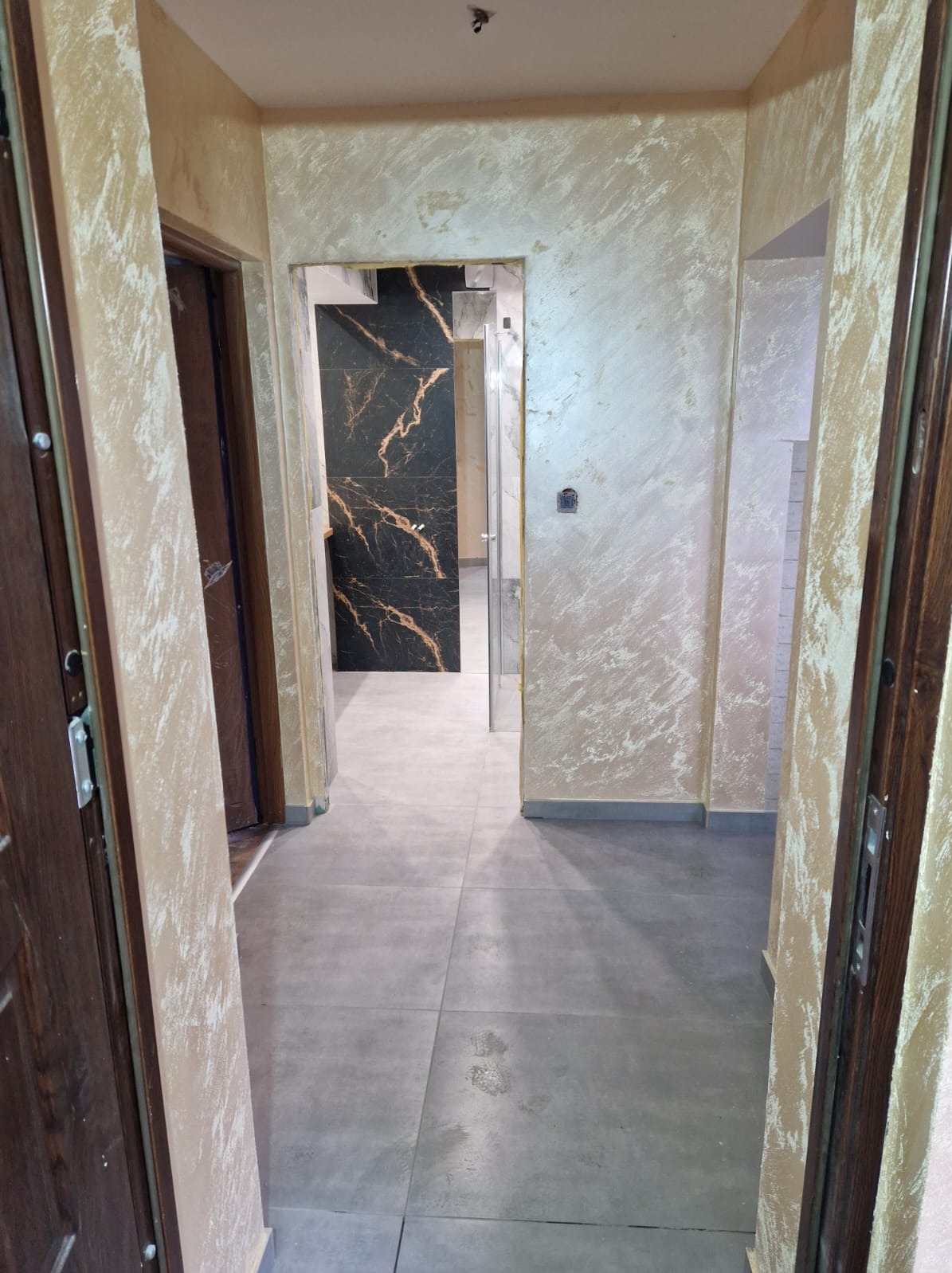 Korytarz po remoncie z szarymi płytkami podłogowymi, ścianami wykończonymi dekoracyjną techniką malarską w odcieniach beżu i złotymi akcentami, widok na łazienkę z czarnymi płytkami imitującymi...