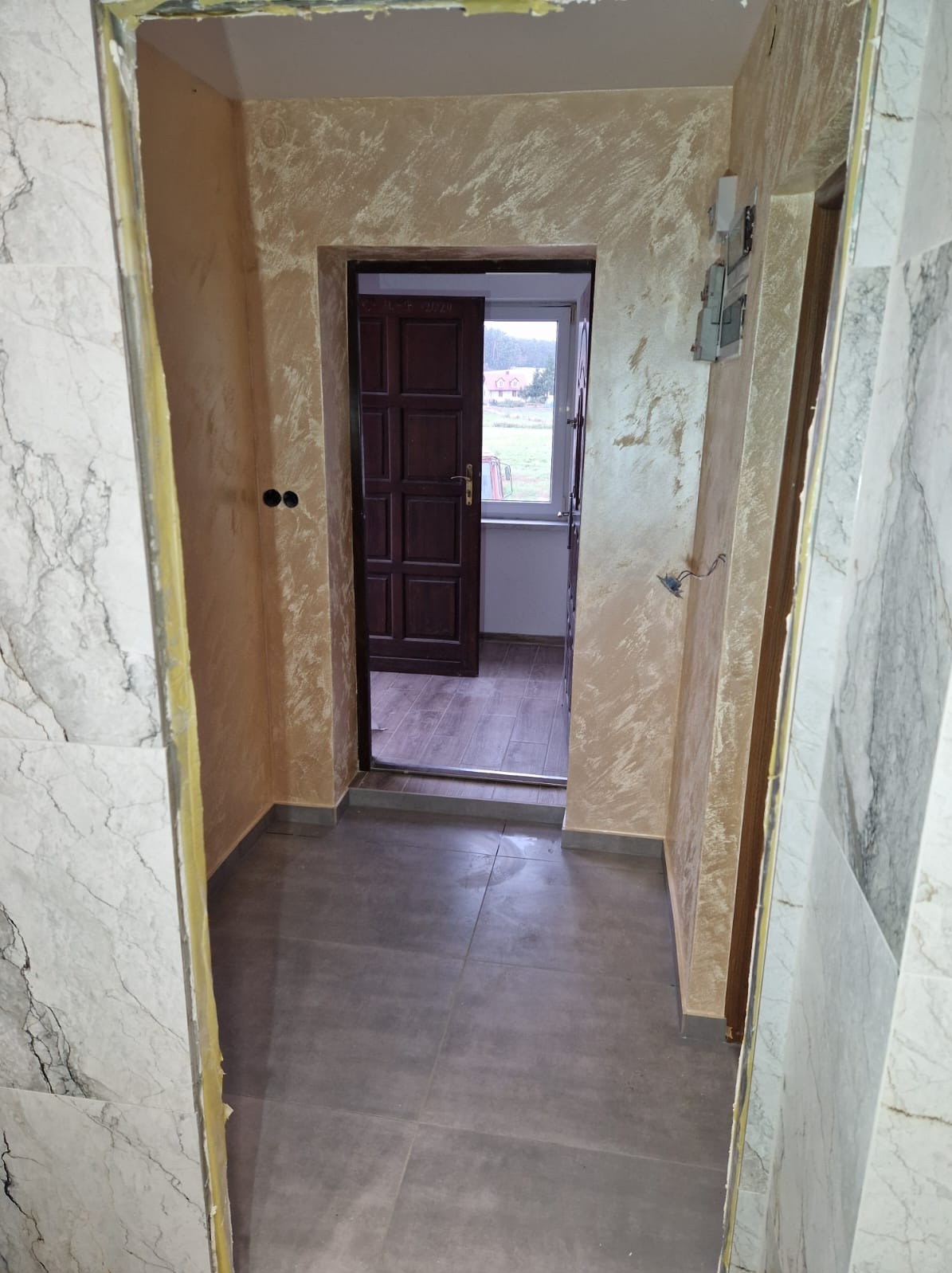 Korytarz po remoncie: szare płytki podłogowe, ściany z efektem dekoracyjnym w odcieniach beżu i brązu, marmurowe wykończenie ścian po lewej stronie, widoczne niedociągnięcia w wykończeniu framugi...