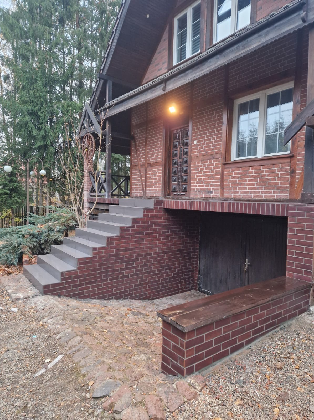 Nowe, szare płytki na schodach wejściowych do domu z czerwonej cegły w Łomży. Widoczny fragment elewacji budynku z drewnianymi elementami i oświetleniem nad drzwiami wejściowymi.