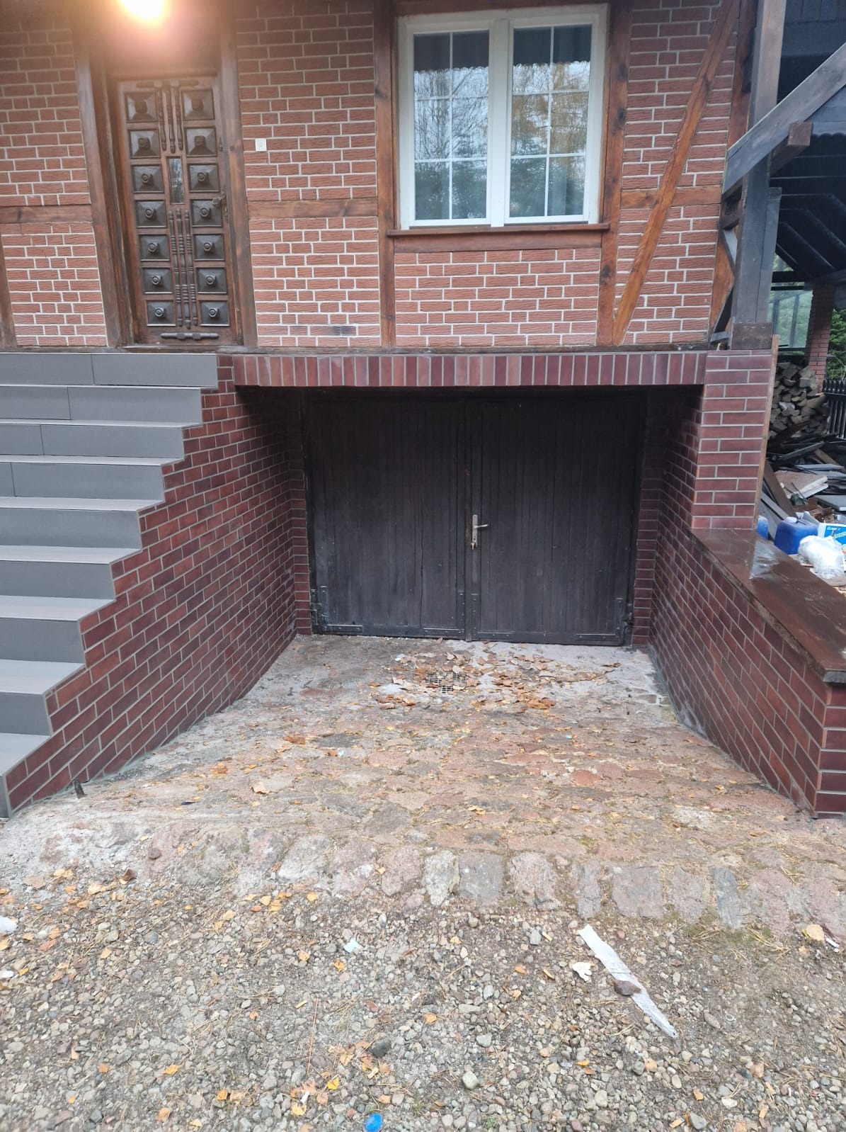 Wykończone schody wejściowe z szarych płytek prowadzące do drzwi domu z ceglaną elewacją i drewnianymi elementami, widoczne również drzwi do piwnicy wykończone cegłą