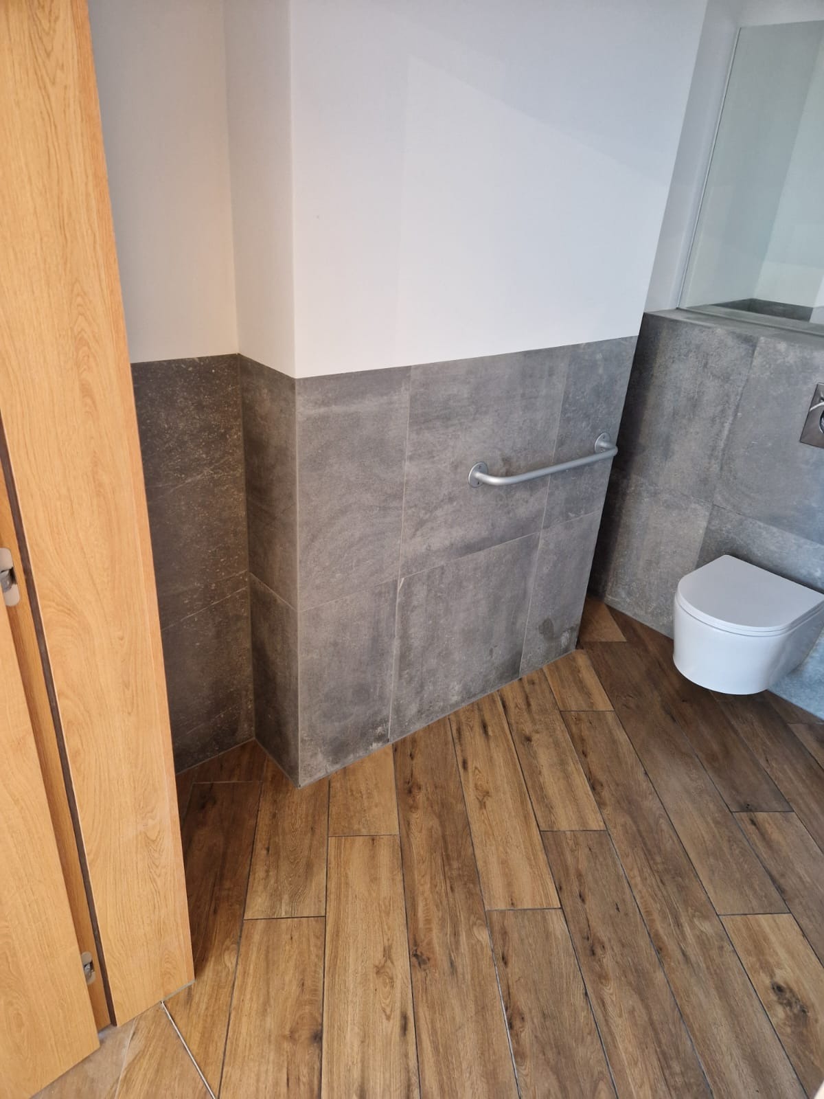 Wykończona łazienka z drewnopodobną podłogą, szarymi płytkami do połowy ściany i białą miską WC. Widoczny fragment drzwi. Nowoczesny design.