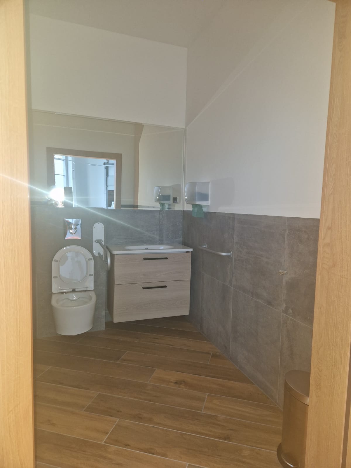 Nowoczesna łazienka po remoncie: drewnopodobne płytki podłogowe, szare płytki ścienne do połowy wysokości, podwieszana szafka z umywalką i lustro.