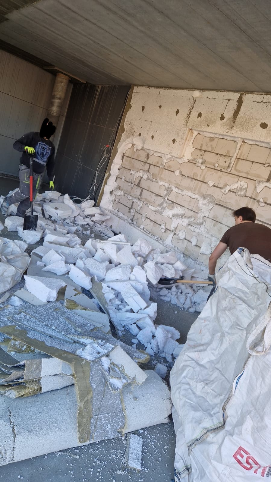 Demontaż ocieplenia: Dwóch robotników usuwa styropian ze ściany, przygotowując podłoże. Fragmenty izolacji i narzędzia na podłodze.