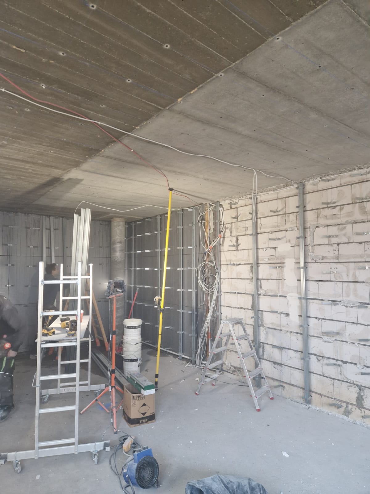Wnętrze w trakcie prac wykończeniowych: widoczne profile na ścianach, instalacja elektryczna, drabiny i narzędzia. Betonowy sufit z widocznymi przewodami.