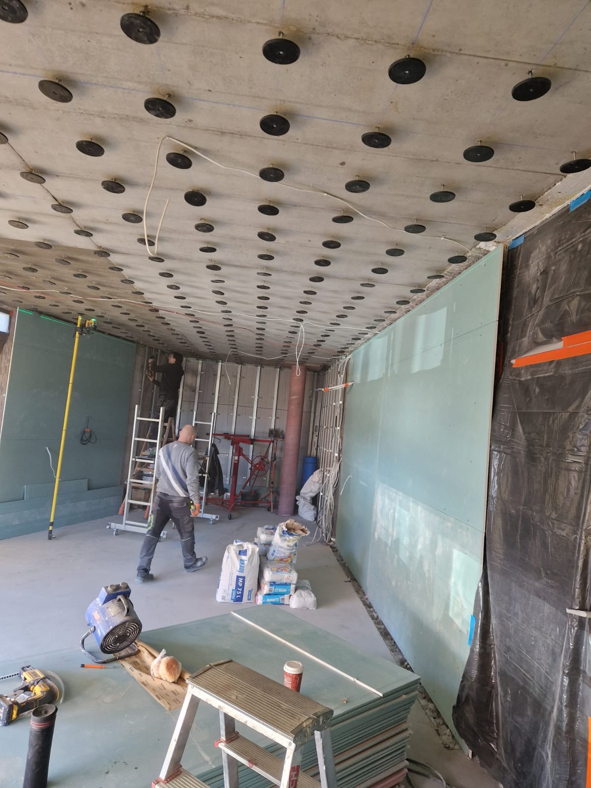 Montaż płyt karton-gips na ścianie i suficie z widocznymi elementami mocującymi. Pracownik na drabinie, narzędzia i materiały budowlane w pomieszczeniu w trakcie prac.