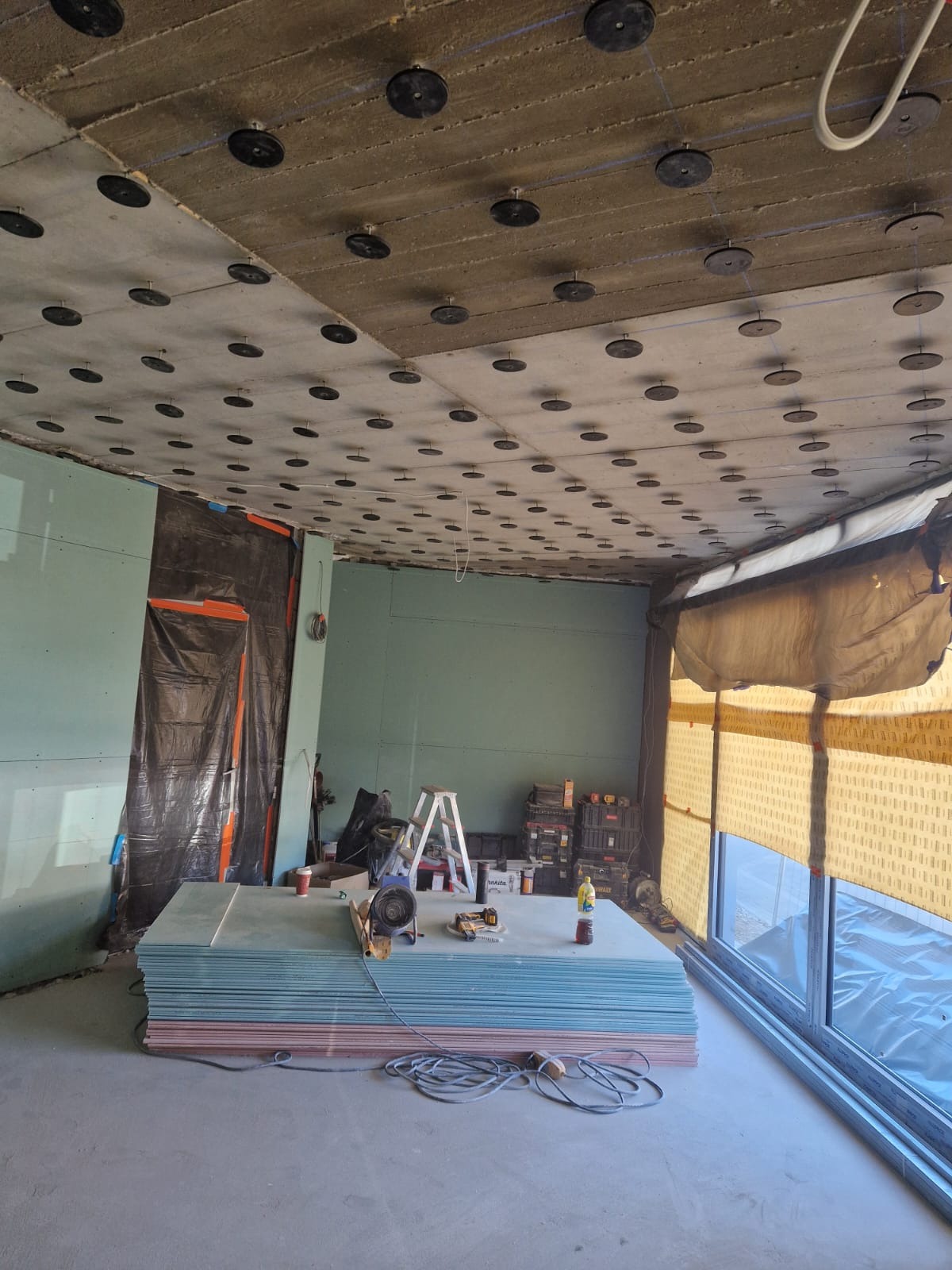 Montaż płyt karton-gips na suficie. Widoczne płyty, narzędzia pracy, drabina, okno. Sufit z elementami mocującymi. Prace wykończeniowe w jasnym pomieszczeniu.